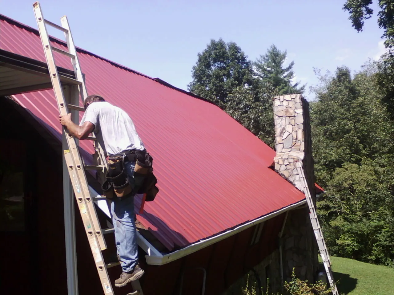 Roofer on a ladder beside a red metal roof and stone chimney — Soffit & Fascia Repair by Roofing Brothers in Swoope
